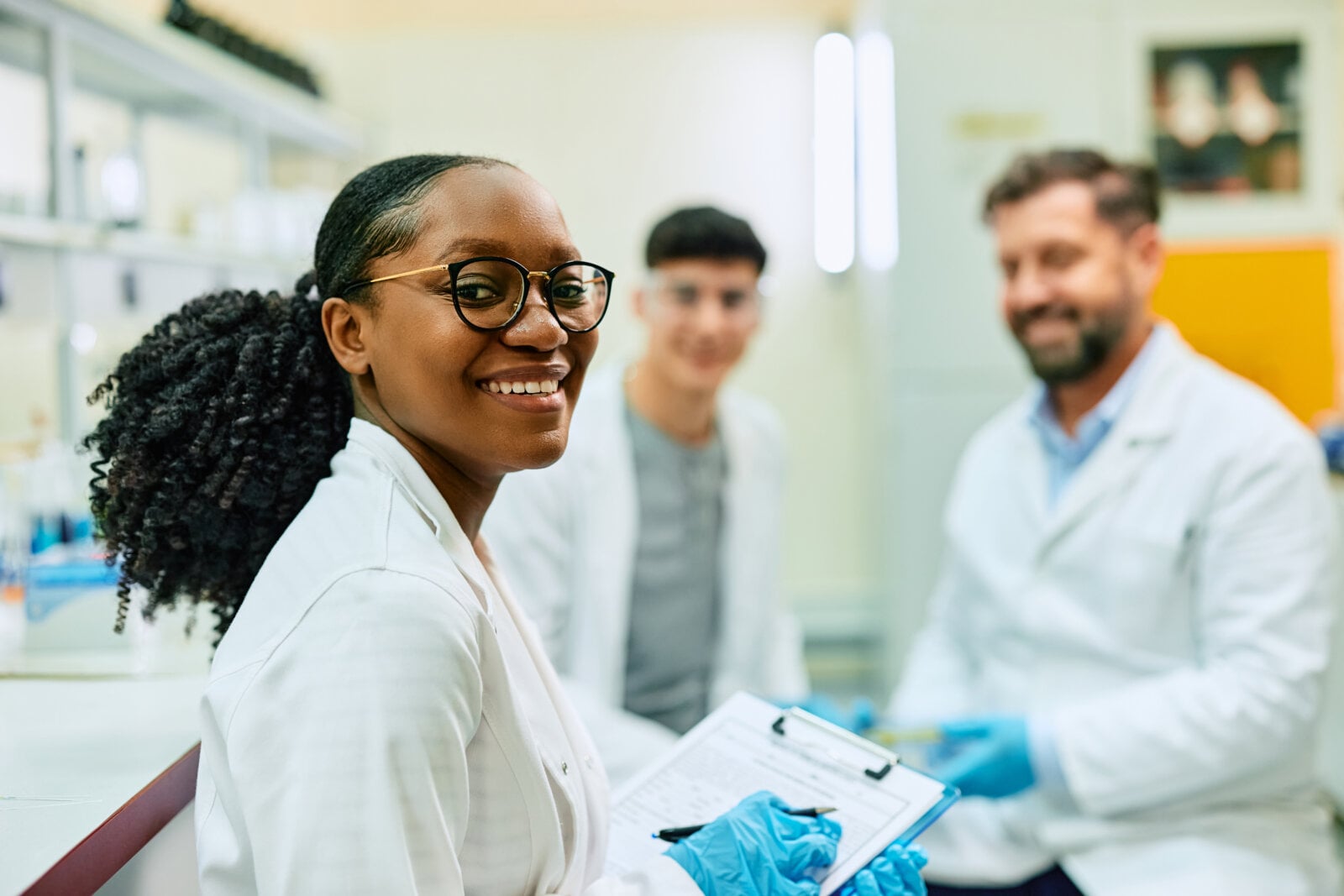 A biochemist holding a clipboard is smiling at the camera in a lab with two of her colleagues.